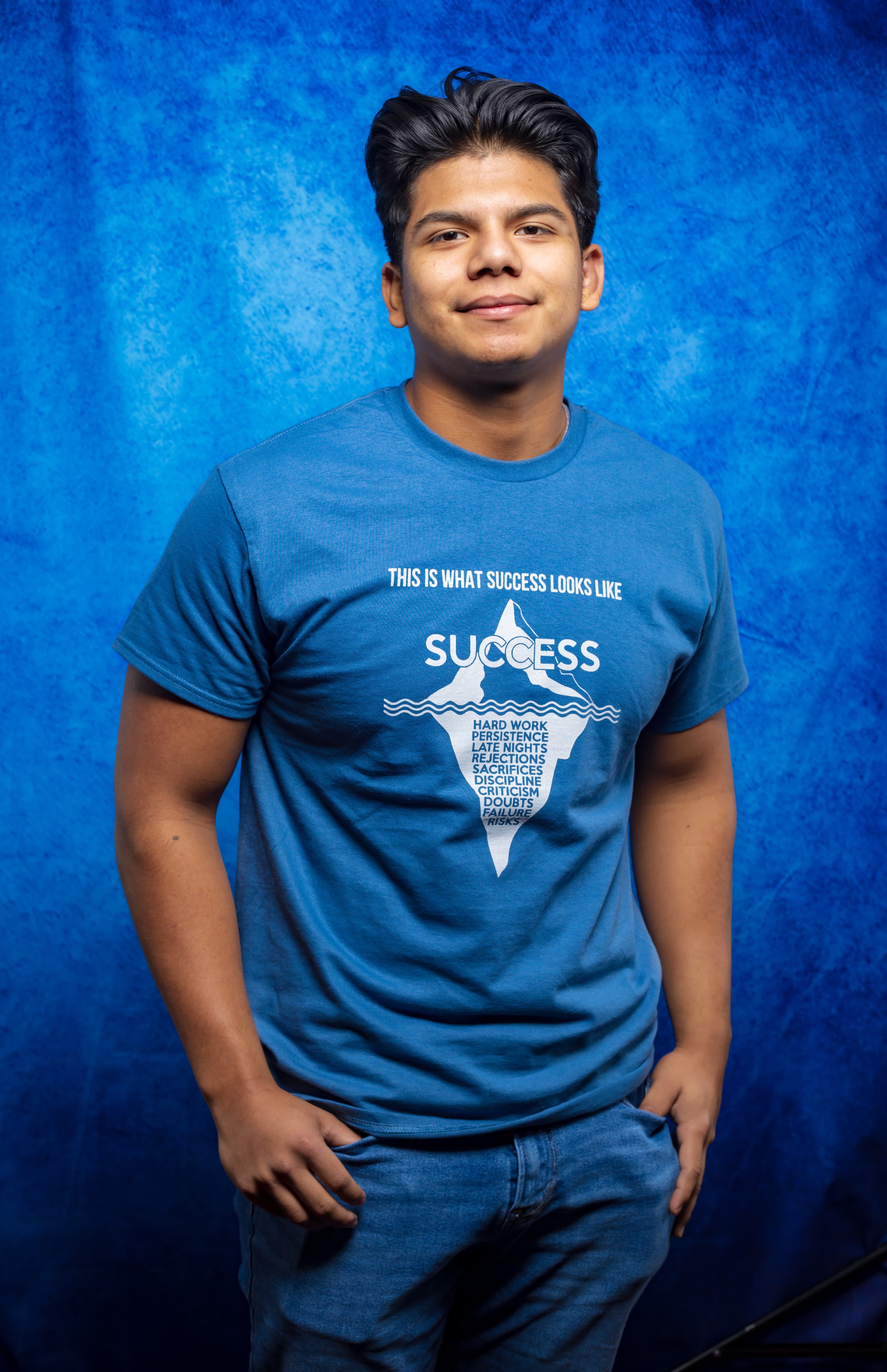 Smiling young man wearing a blue success iceberg graphic t-shirt against a textured blue backdrop.