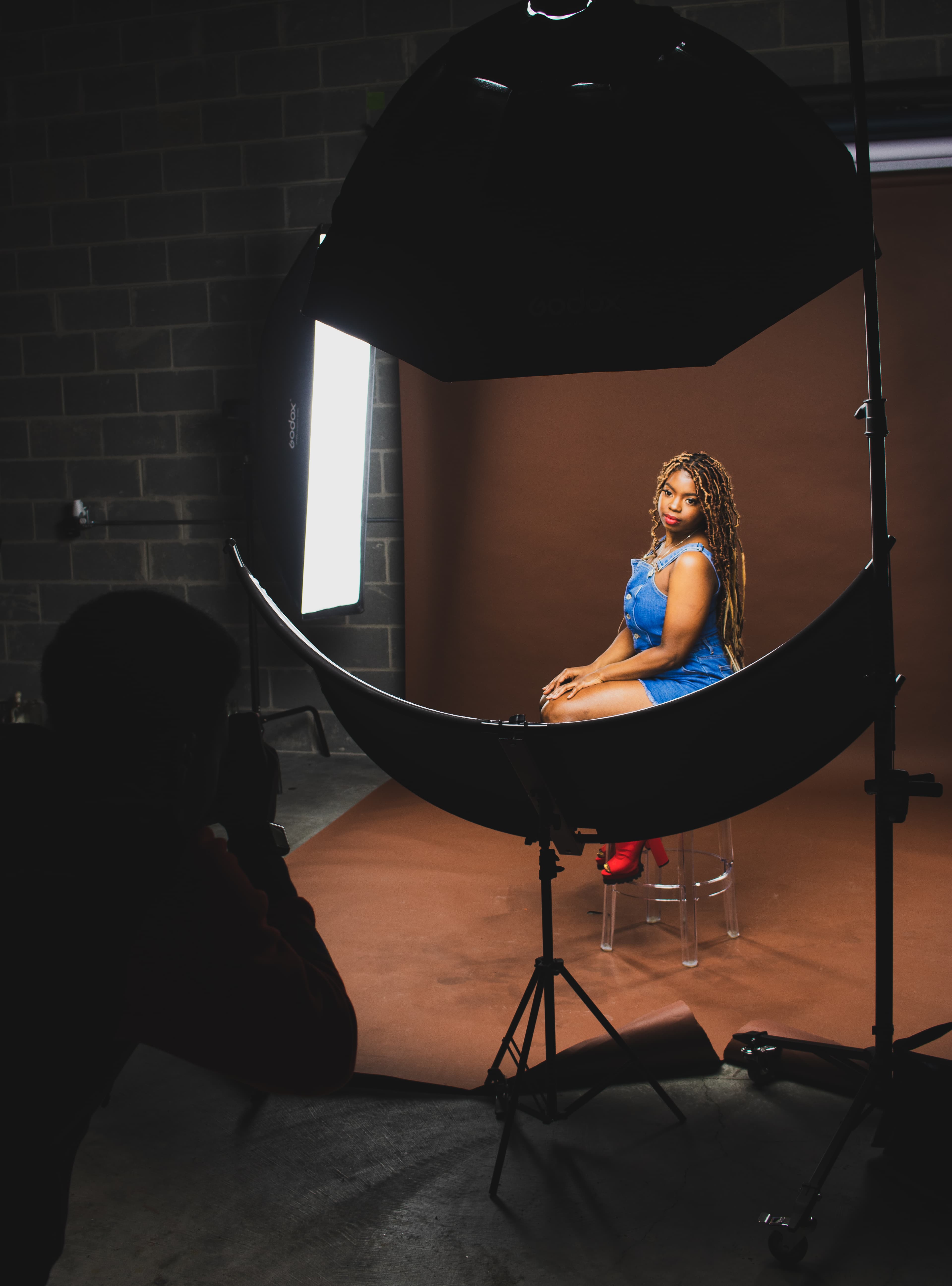 Woman in denim romper poses for a photographer in a studio with professional lighting equipment.