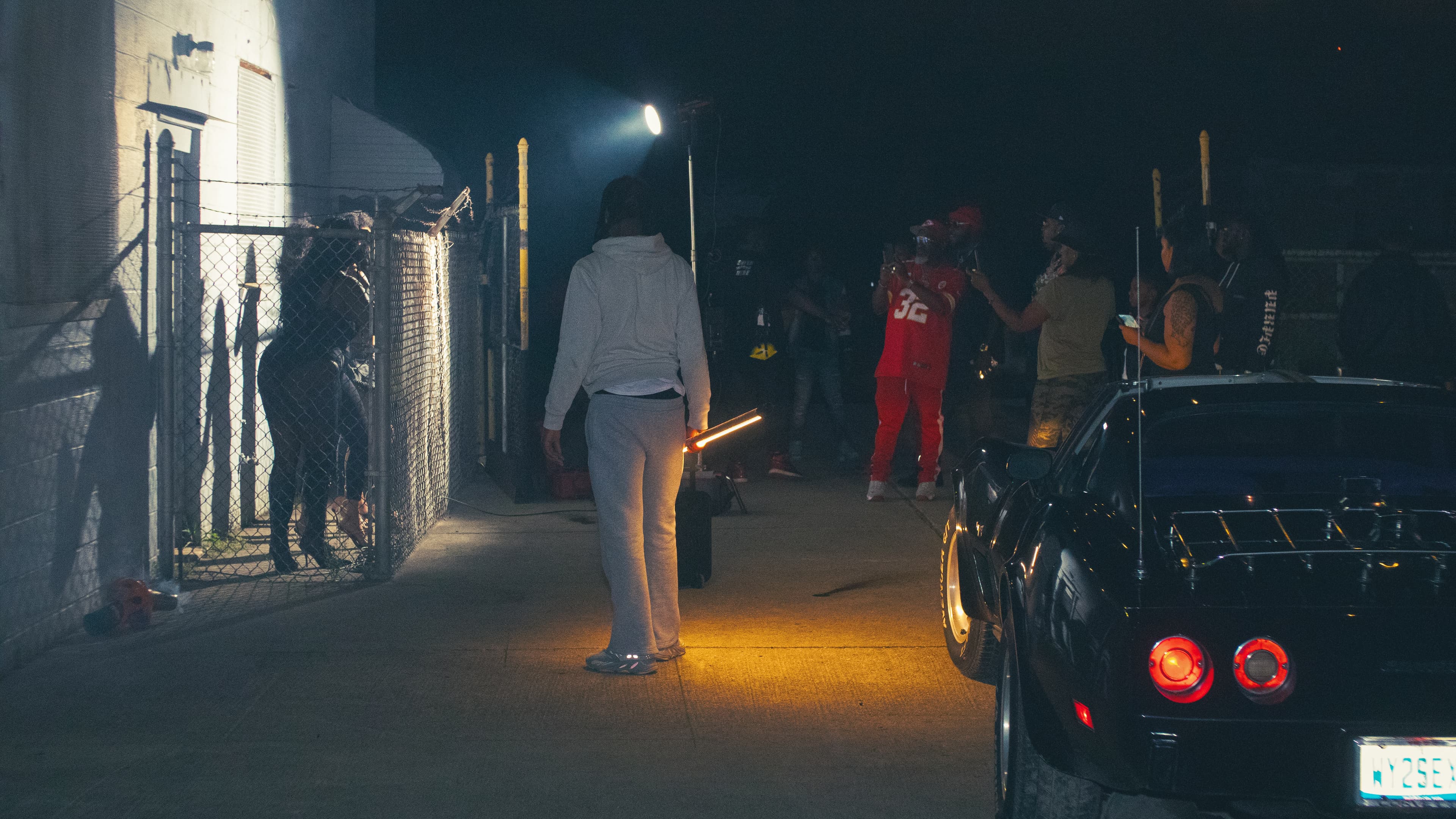 People gather on a nighttime film set featuring a black classic car and bright spotlights.