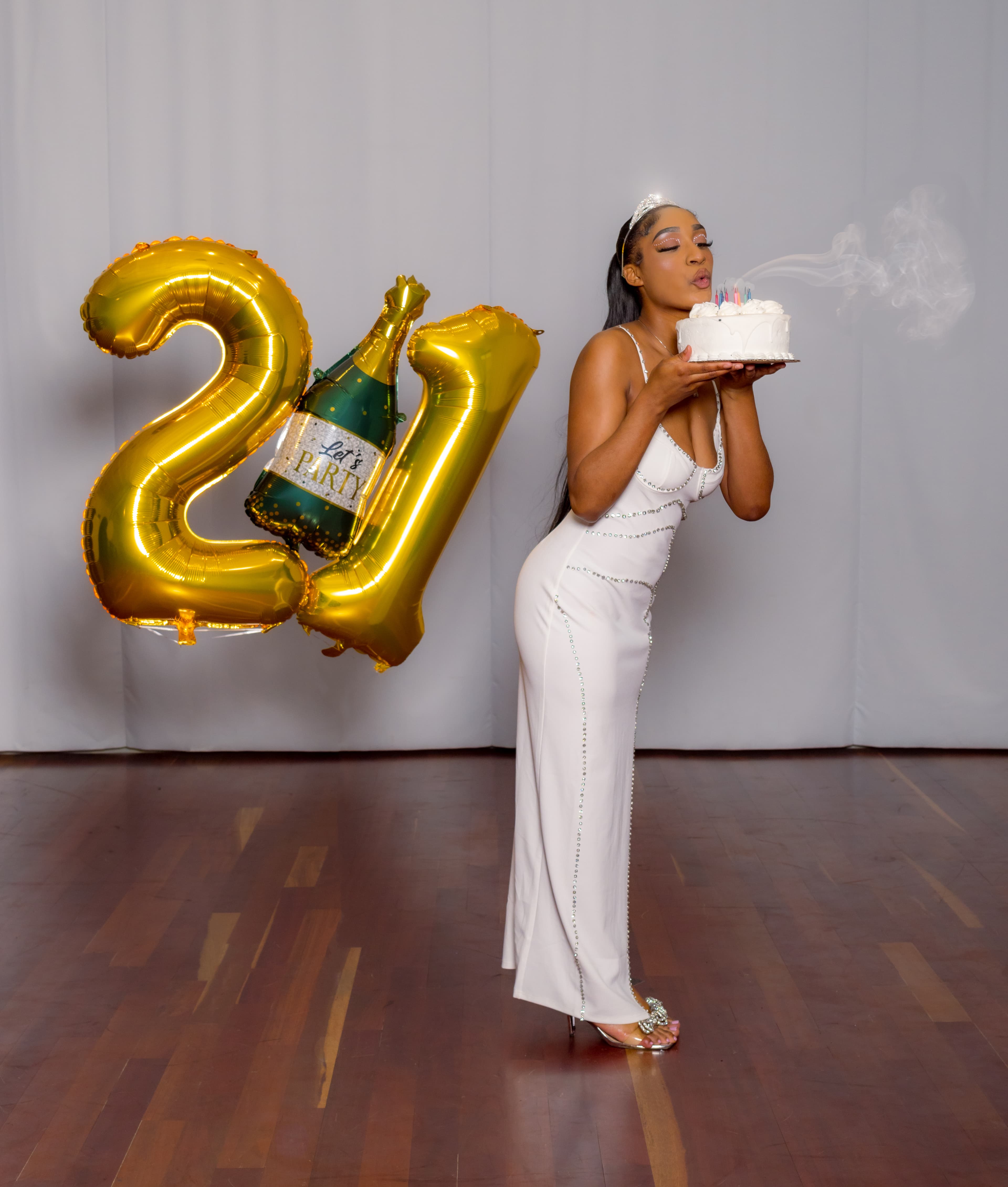 Woman in white dress blowing out candles on cake beside large gold 21 balloons.